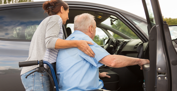 A lady helping an elder male getting into a car from his wheelchair