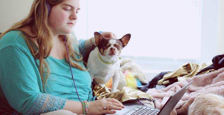 Young female sitting on her bed with her dog and using her laptop