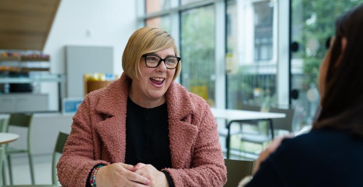 A lady in a pink jacket is sitting at a table talking to another person