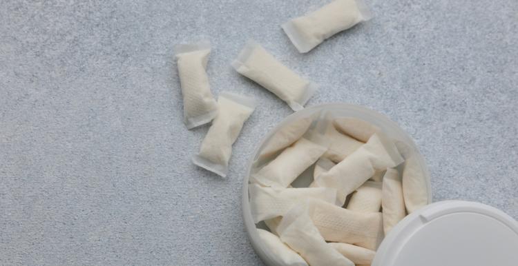 A white container holds nicotine pouches on a light gray surface. Several pouches are placed outside the container. The scene shows the pouches arranged casually.
