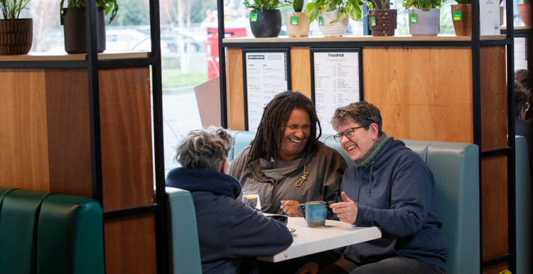 3 adults sitting in a cafe with hot drinks while chatting and laughing