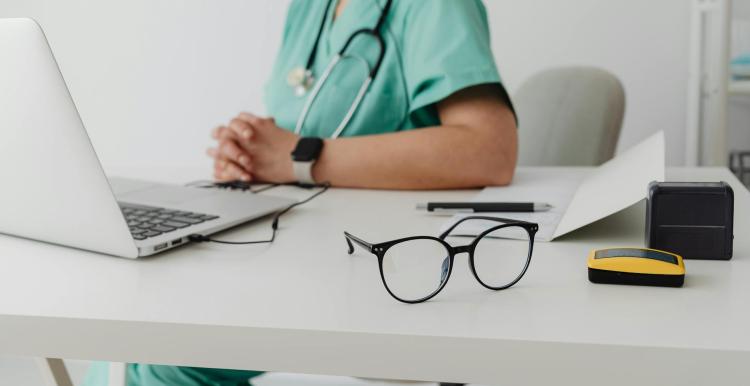 View of a desk with a laptop, glasses and a doctor sitting on the other side