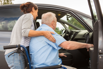 A lady helping an elder male getting into a car from his wheelchair