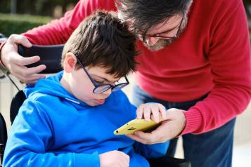 A male helping a young child in a wheelchair with his mobile phone