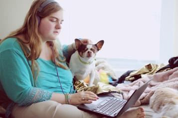 Young female sitting on her bed with her dog and using her laptop