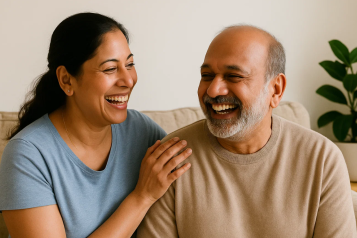 An older daughter sitting next to her elderly father on a couch, both laughing and smiling.
