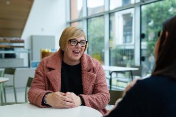 A lady in a pink jacket is sitting at a table talking to another person