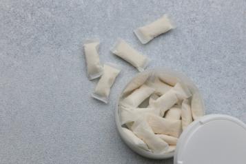 A white container holds nicotine pouches on a light gray surface. Several pouches are placed outside the container. The scene shows the pouches arranged casually.