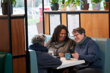 3 adults sitting in a cafe with hot drinks while chatting and laughing