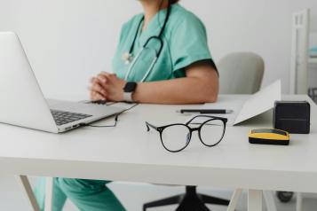 View of a desk with a laptop, glasses and a doctor sitting on the other side
