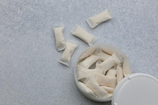 A white container holds nicotine pouches on a light gray surface. Several pouches are placed outside the container. The scene shows the pouches arranged casually.