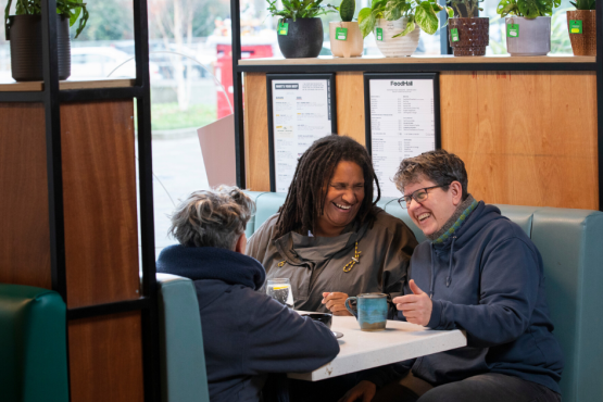 3 adults sitting in a cafe with hot drinks while chatting and laughing