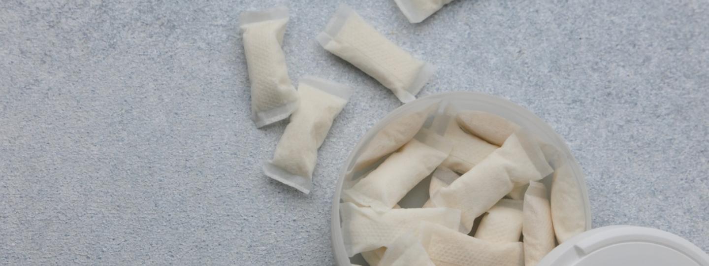 A white container holds nicotine pouches on a light gray surface. Several pouches are placed outside the container. The scene shows the pouches arranged casually.